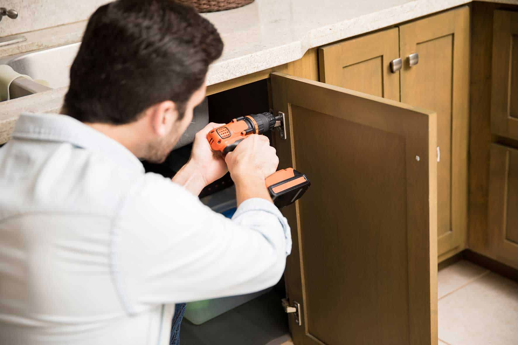 young-handyman-using-power-drill-fix-door-kitchen-cabinet-(1)